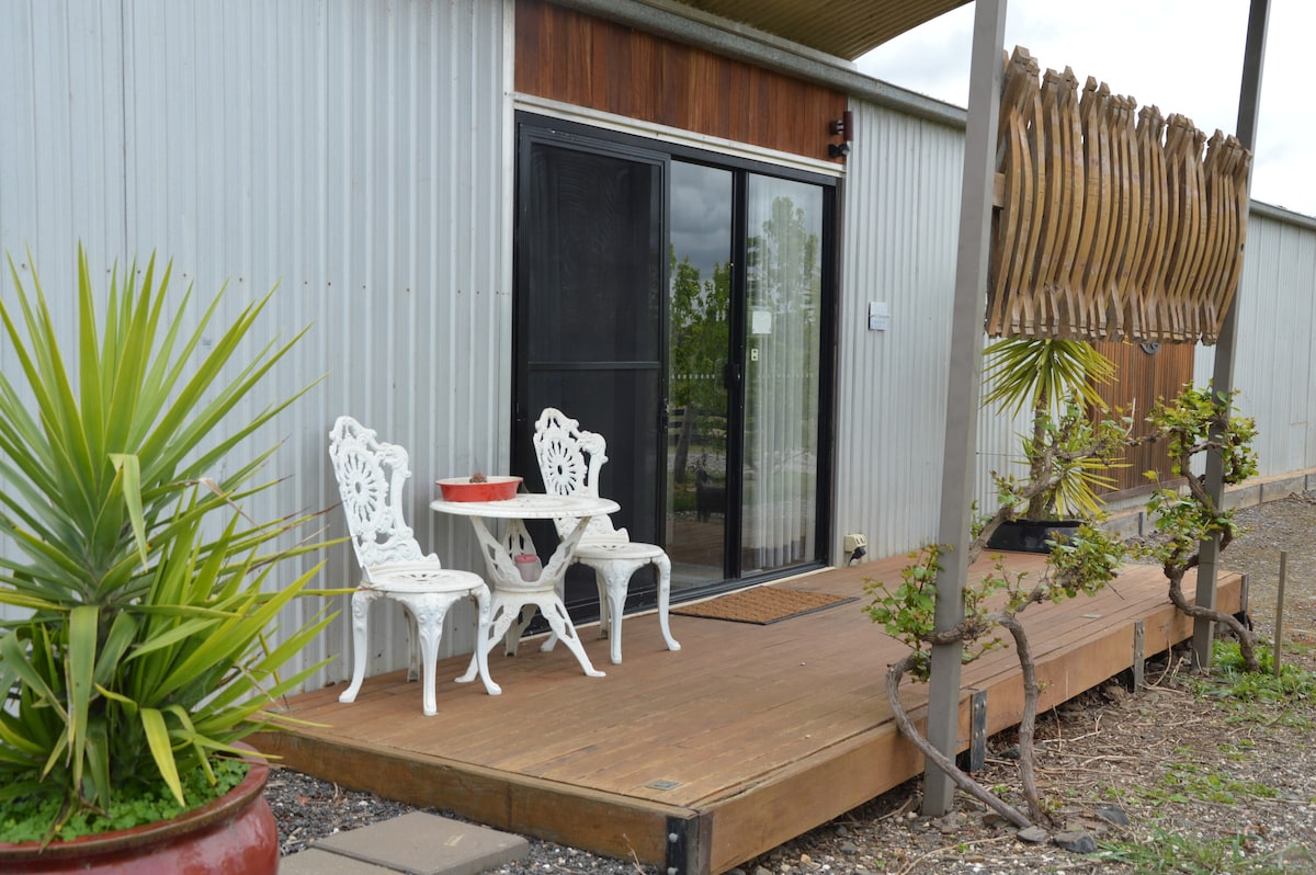 A wooden deck area is featured outside the studio, with two white chairs and a small table positioned for relaxation. Nearby, a green plant adds a touch of nature, while unique wooden decor hangs decoratively beside the door.