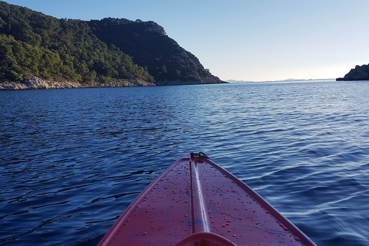 A red kayak is positioned in the foreground, with calm blue waters extending towards a green hillside in the background. The surface of the water reflects the soft morning light, while distant islands can be faintly seen on the horizon.