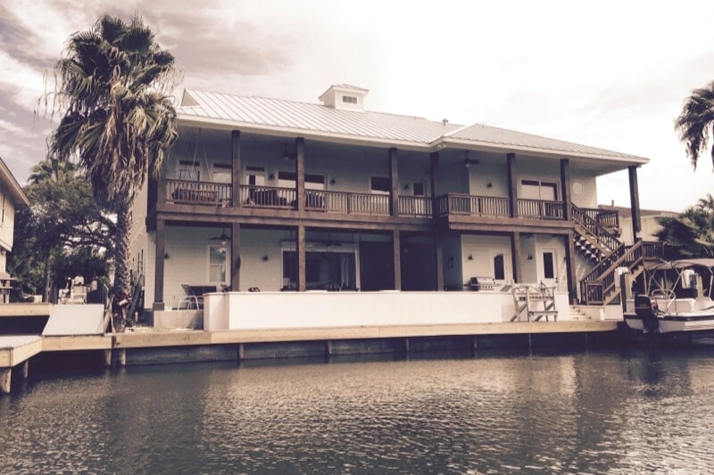 A two-story canal front home features a spacious deck with multiple outdoor seating areas. The exterior showcases a light-colored façade with large windows. A boat dock is visible, alongside palm trees framing the serene waterway.