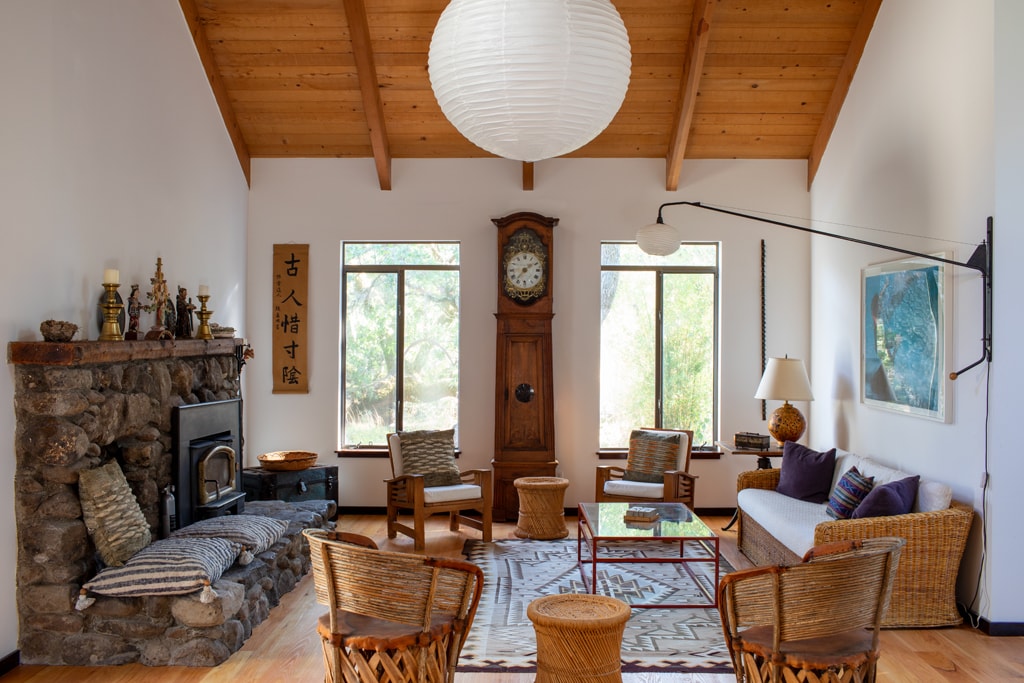 A spacious living room features high ceilings with exposed wooden beams. A large stone fireplace is surrounded by comfortable seating, including wicker chairs and a cozy sofa. Natural light filters through large windows, illuminating the room's warm wood accents and calming decor elements.