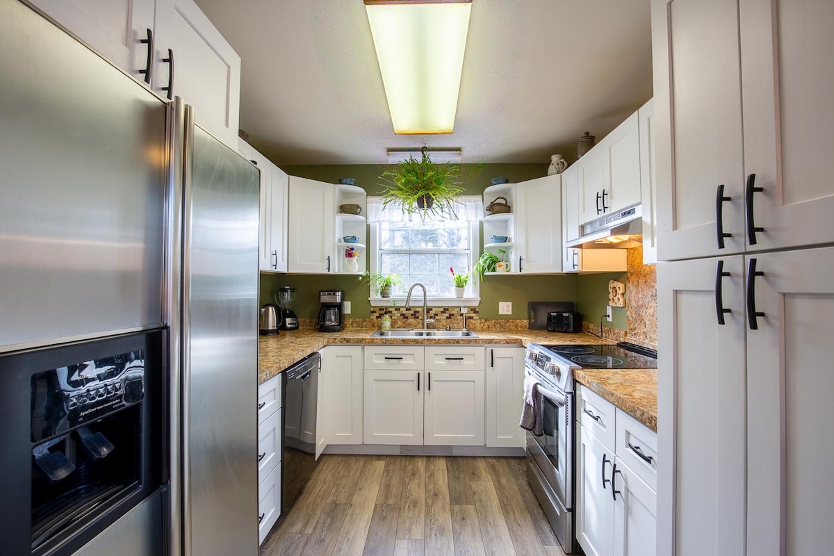 The kitchen features modern stainless steel appliances, including a refrigerator and oven, complemented by white cabinetry and granite countertops. A spacious layout allows for easy movement, and natural light filters in through a window adorned with potted plants.