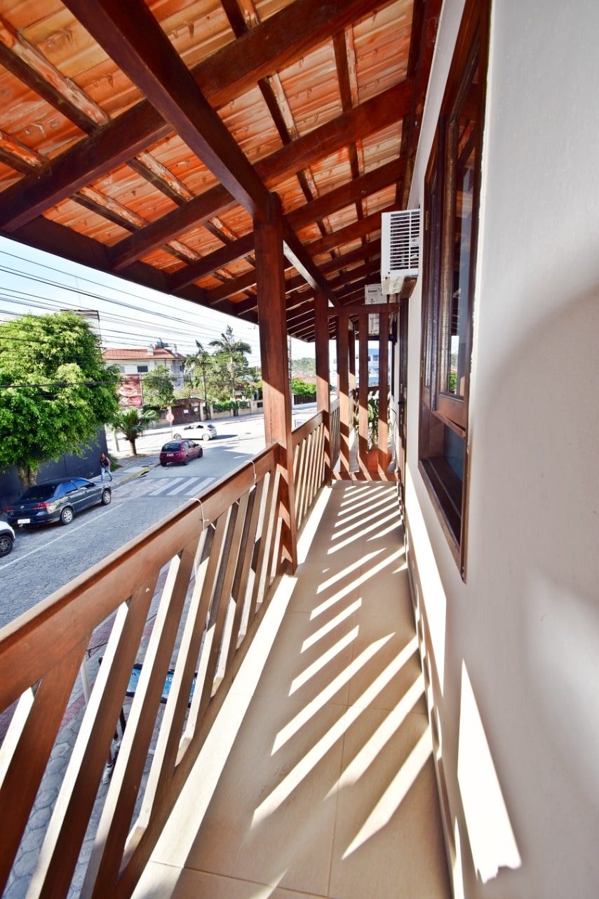 A covered balcony with wooden railings is featured, providing ample space for relaxation. Natural light creates shadows on the tiled floor, enhancing the openness of the area. Views of the surrounding street and greenery are visible, providing a connection to the vibrant outdoor environment.