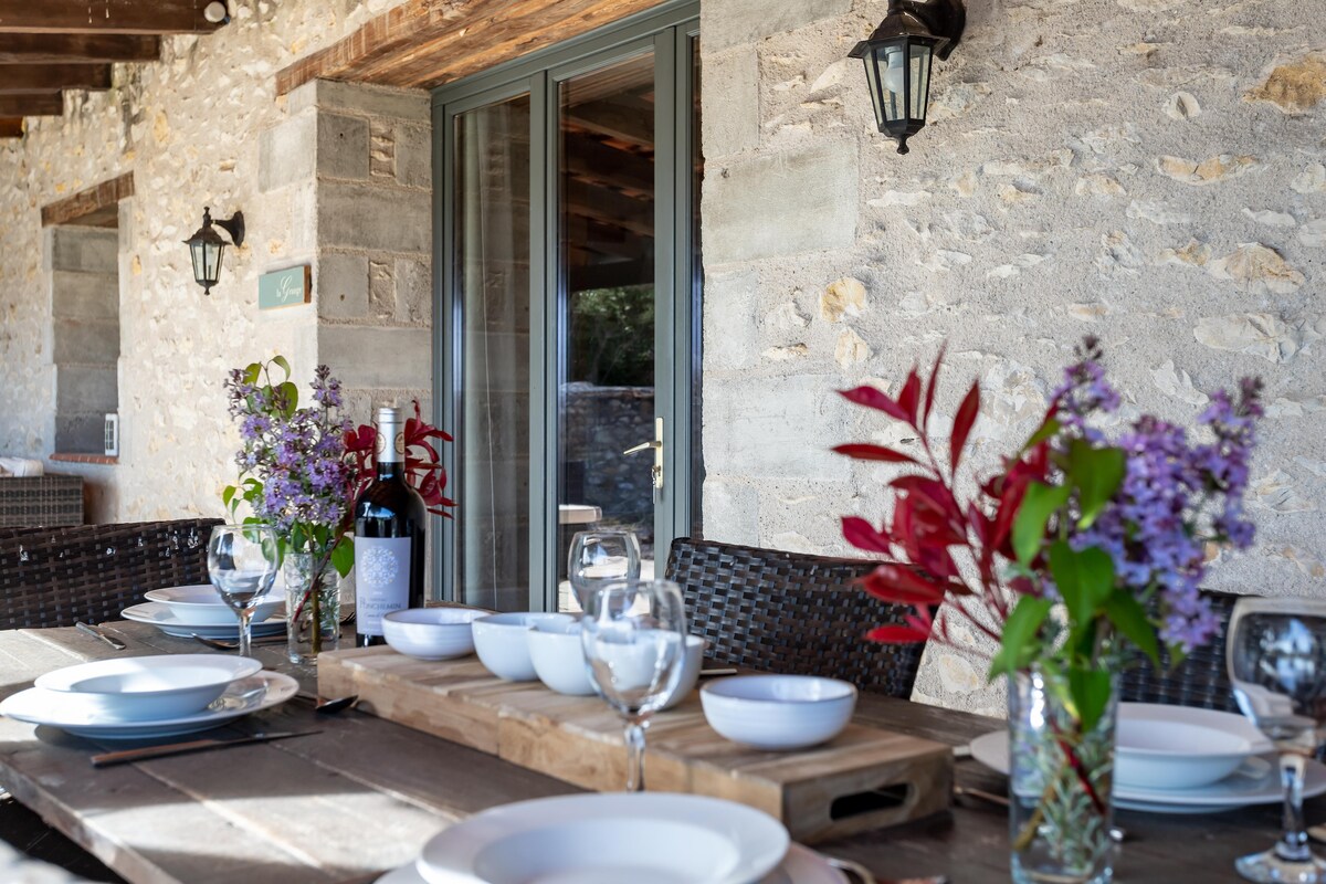 A rustic dining table is set outside, featuring white plates and glasses arranged neatly. A bottle of wine stands amongst vibrant flowers in glass vases, enhancing the natural ambiance of the covered terrace. Stone walls and soft lighting create a welcoming space.