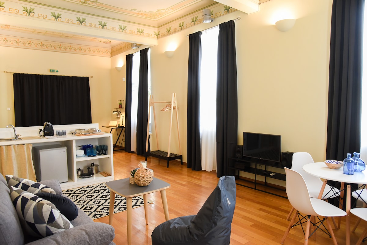 An open-plan studio space showcases high ceilings adorned with intricate designs. Natural light enters through large windows, illuminating the elegant furnishings. A cozy seating area features a bean bag chair and a patterned rug, complemented by a compact kitchenette and a dining table set for four.