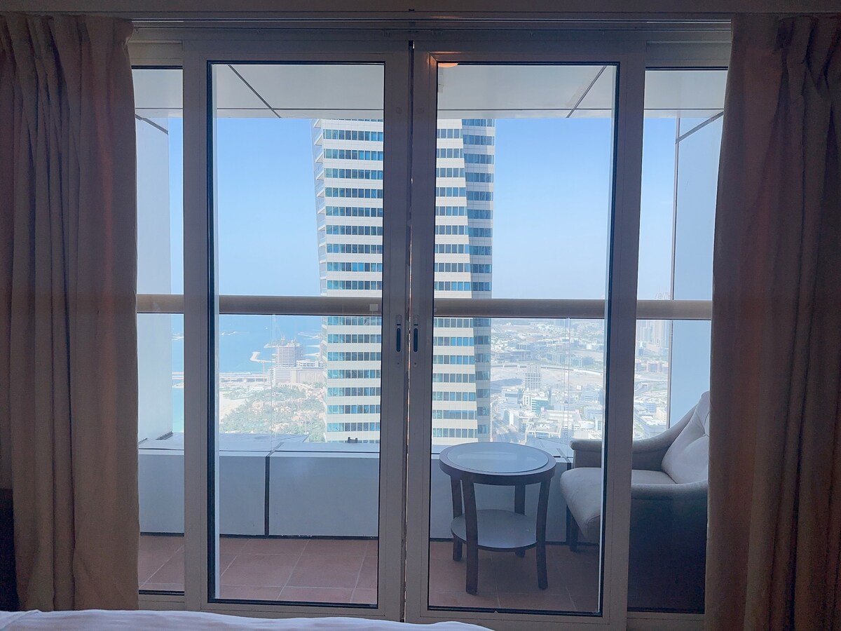 A view from the private room showcases large glass sliding doors that lead to a balcony. A small round table and a chair are visible on the balcony, with a striking urban landscape and partial sea view beyond.