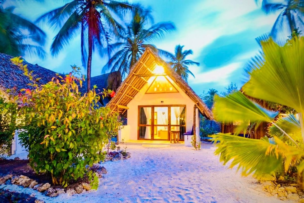 A charming bungalow features a thatched roof and large glass doors, allowing natural light to fill the interior. Surrounding palms and lush greenery create a tropical ambiance, while sandy pathways lead to the entrance, adorned with vibrant plants.