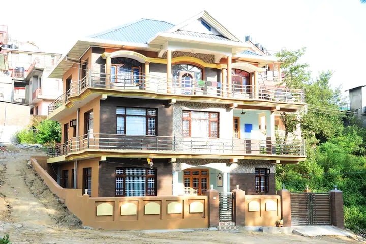 The three-story house features a covered entrance with columns and a balcony on each level. Large windows provide natural light, while a blend of brick and wood accents creates a welcoming façade. The surrounding area is peaceful, with greenery visible in the background.