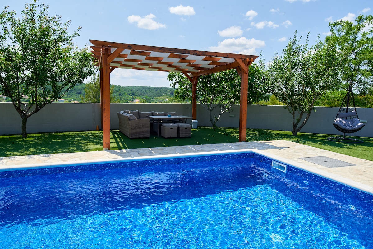 A private pool with a vibrant blue surface is set against a backdrop of green trees and lush grass. A shaded seating area with a modern couch is positioned nearby, offering a relaxing space. The overall atmosphere is tranquil and inviting, perfect for families.