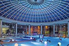 A spacious indoor pool area features a striking glass dome ceiling, allowing natural light to illuminate the water. Softly colored lights float in the pool, and guests can be seen enjoying the tranquil atmosphere. The surrounding walls are adorned with large windows, enhancing the openness.