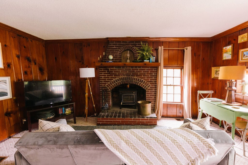 A warm living area showcases rich wooden wall paneling and features a brick fireplace at its center. A comfortable sofa is positioned in the foreground, with a patterned throw draped casually over it. A television and various decorative elements complete the inviting space.