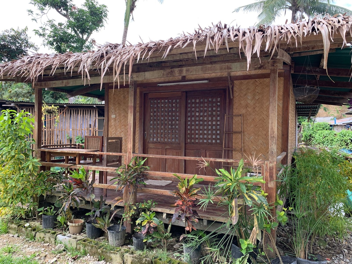 A rustic wooden cottage is surrounded by lush greenery and potted plants. The structure features a thatched roof and a spacious porch with multiple chairs, showcasing a welcoming entrance with intricately designed wooden doors.