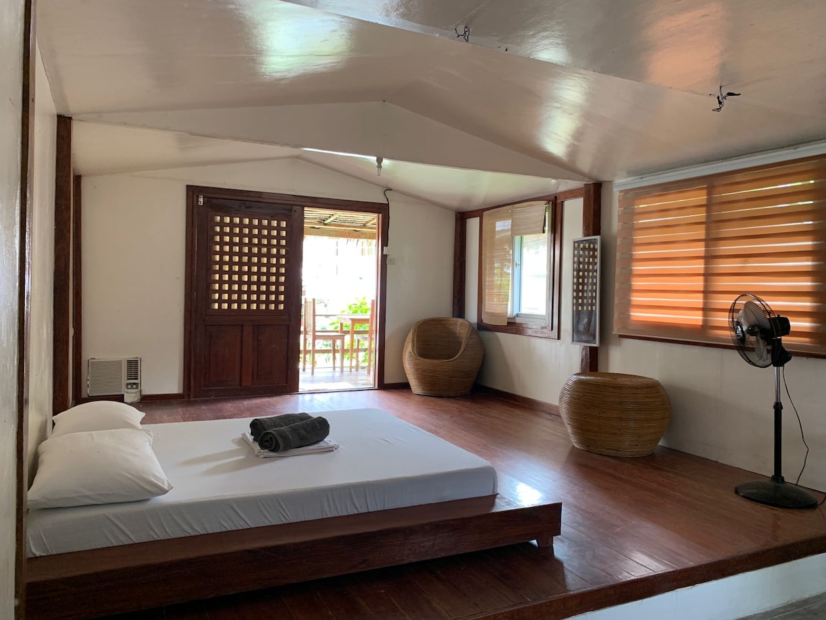 A spacious room features polished wooden flooring and a large bed with a white mattress. Natural light enters through a window with wooden blinds. Two wicker chairs are placed in one corner, and a standing fan provides ventilation. The entrance includes a wooden door with a lattice design.