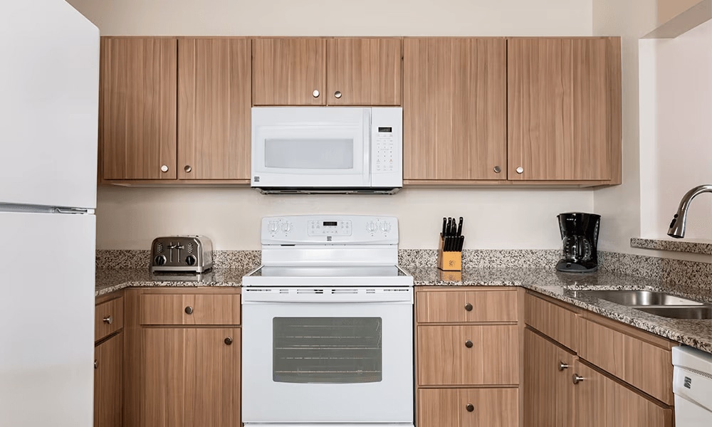 A full kitchen is equipped with modern appliances, including a refrigerator, oven, microwave, and coffee maker. The countertops feature a speckled granite design with wooden cabinetry providing ample storage. Kitchen utensils are neatly arranged next to the sink.