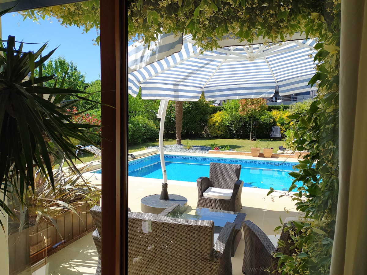 A view of the outdoor area features a spacious terrace overlooking a heated pool. Sun loungers and a striped umbrella provide shade, while potted plants and greenery surround the space, creating a serene atmosphere for relaxation.