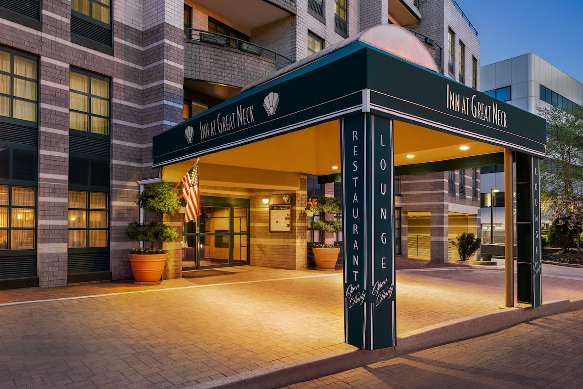 A welcoming entrance is highlighted by a covered awning, featuring the establishment's name prominently displayed. Potted plants add greenery along the pathway, while an American flag gently waves nearby, indicating the welcoming atmosphere of the entrance to the hotel.
