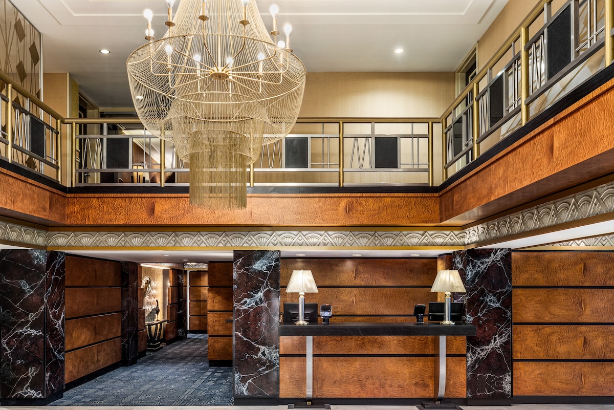 A grand lobby area is highlighted by a large chandelier suspended from the ceiling. Dark wooden panels and marble accents create a sophisticated atmosphere. A reception desk is positioned in the foreground, while a staircase is visible in the background, providing access to the upper level.