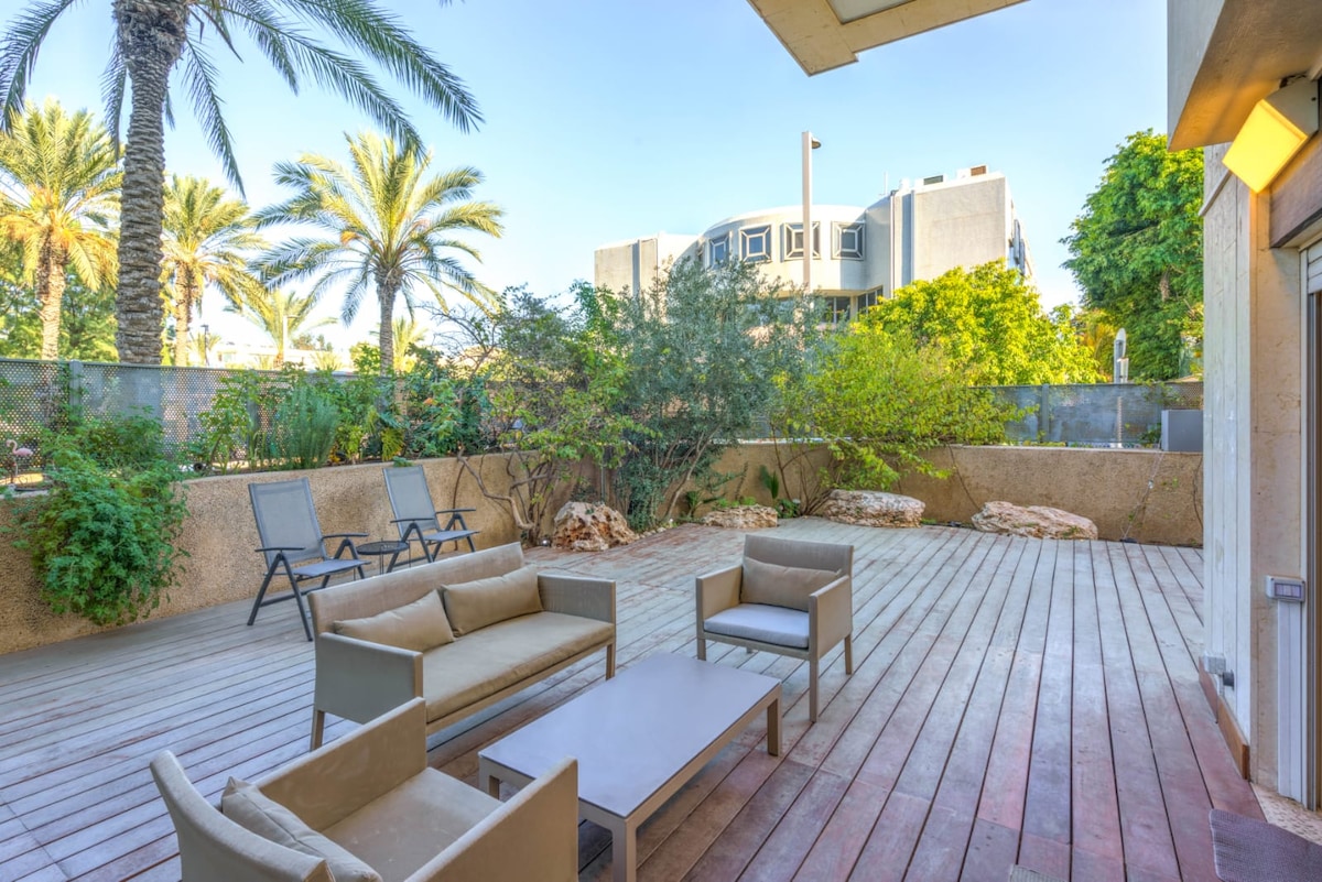 A spacious terrace features a wooden deck surrounded by lush greenery, including palm trees and shrubs. Comfortable seating is provided with a modern outdoor table and lounge chairs, creating an inviting space for relaxation and enjoyment of the natural surroundings.