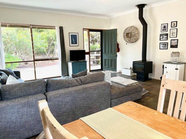 An open-plan living area features a large, comfortable sectional sofa and a cozy wood-burning stove. A dining table sits adjacent to the kitchen, and dual doors open to a natural view outside, inviting sunlight into the space.