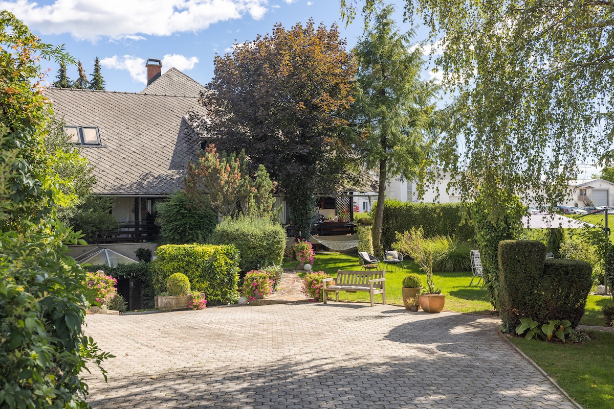 The garden pathway leads to a charming home surrounded by lush greenery and floral arrangements. A wooden bench is positioned among the vibrant plants, providing a serene spot for relaxation. The landscaping features well-trimmed hedges and trees, contributing to a tranquil outdoor setting.