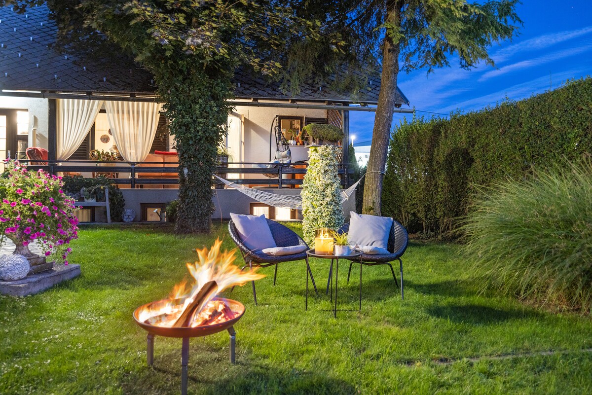 A serene garden area is depicted with a fire pit surrounded by two chairs on a grassy lawn. Colorful flowers are positioned nearby, and a shaded patio can be seen in the background. Soft evening light creates a calm and inviting ambiance.