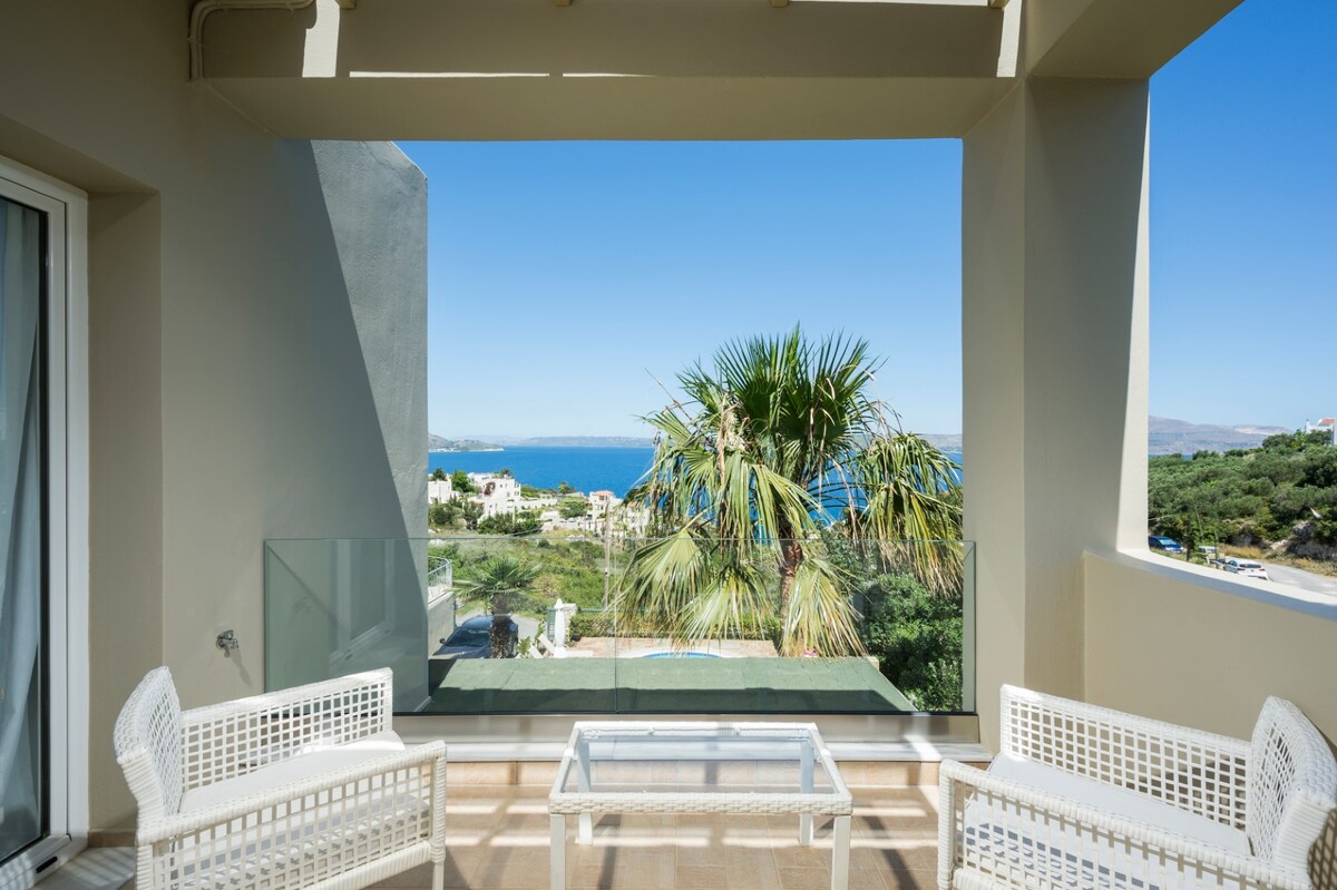 A spacious outdoor veranda is shown, featuring two white wicker chairs and a glass table. Lush greenery and a palm tree frame the view of the azure sea and landscape beyond. The clear sky adds to the serene atmosphere of the space.
