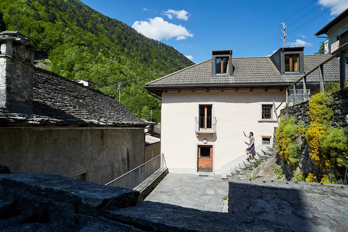 The exterior of the renovated house is presented, showcasing a two-story structure with a light-colored façade. Stairs lead to the main entrance, framed by a stone pathway. Surrounding green hills and trees enhance the natural setting, while a blue sky adds brightness.
