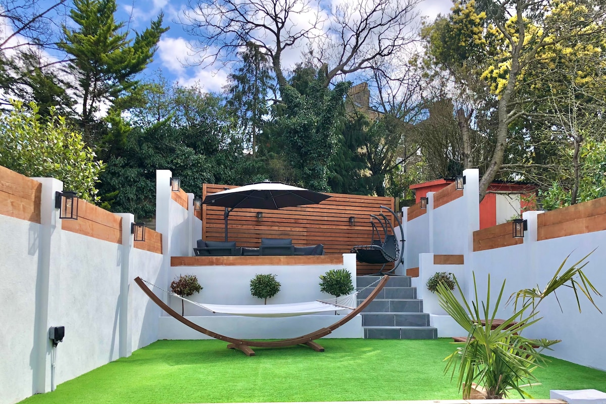 A spacious Mediterranean-style garden is visible, featuring a green lawn, a hammock, and comfortable seating under a large umbrella. Surrounding walls are adorned with plants, providing a serene atmosphere. Steps lead to a raised wooden deck, enhancing the outdoor space.