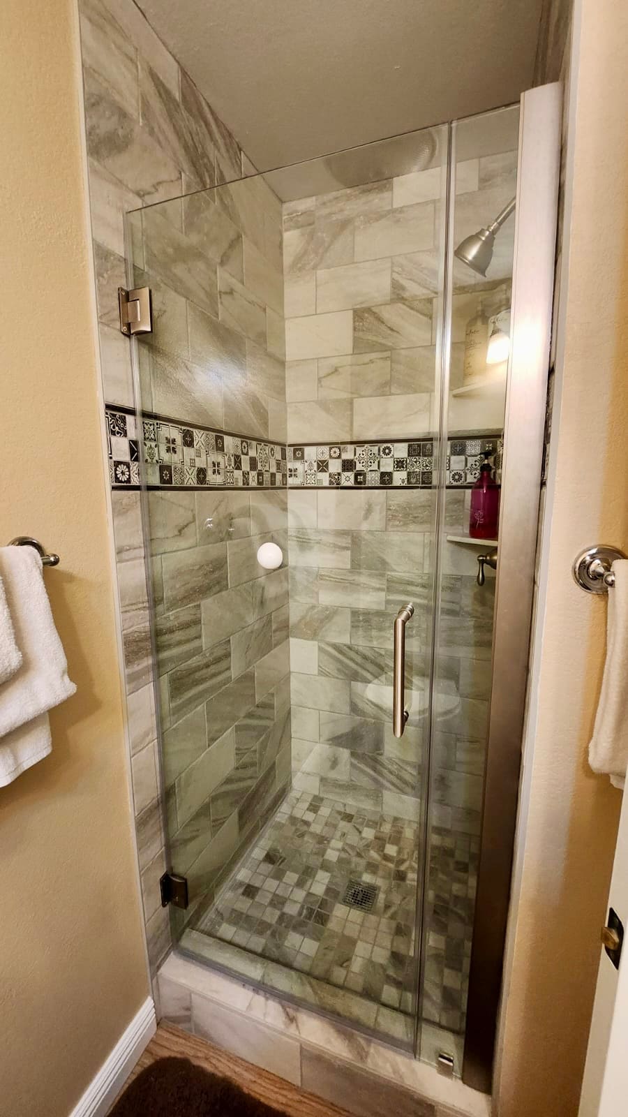 A modern glass shower enclosure is presented, featuring sleek gray and white marbled tiles. The shower head is mounted above, with a mosaic floor of small square tiles. Towels are neatly hung nearby, complementing the contemporary style.