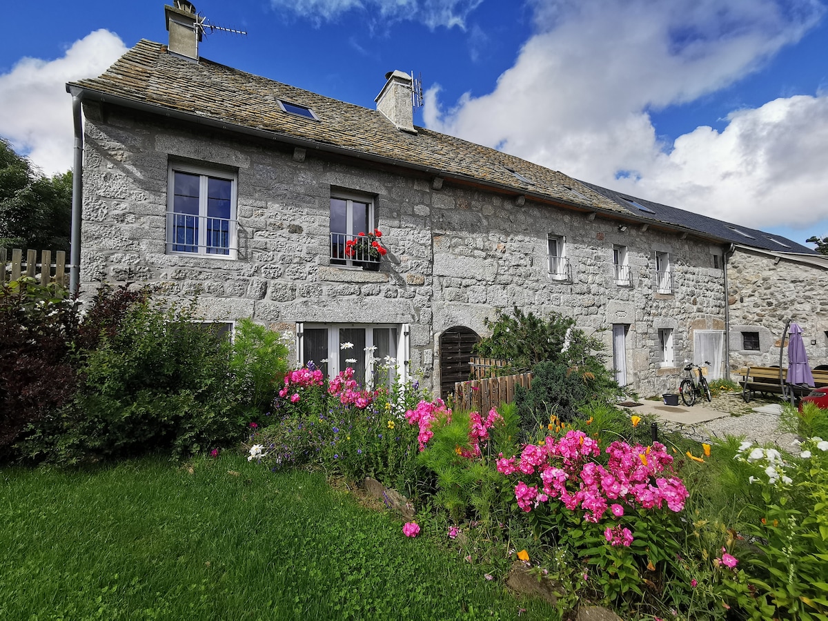 The exterior of the gîte presents a charming stone building with a flower-filled garden in the foreground. A colorful array of flowers, including pink blooms, enhances the outdoor space. Bright windows and a small balcony adorned with potted flowers contribute to the inviting appearance.