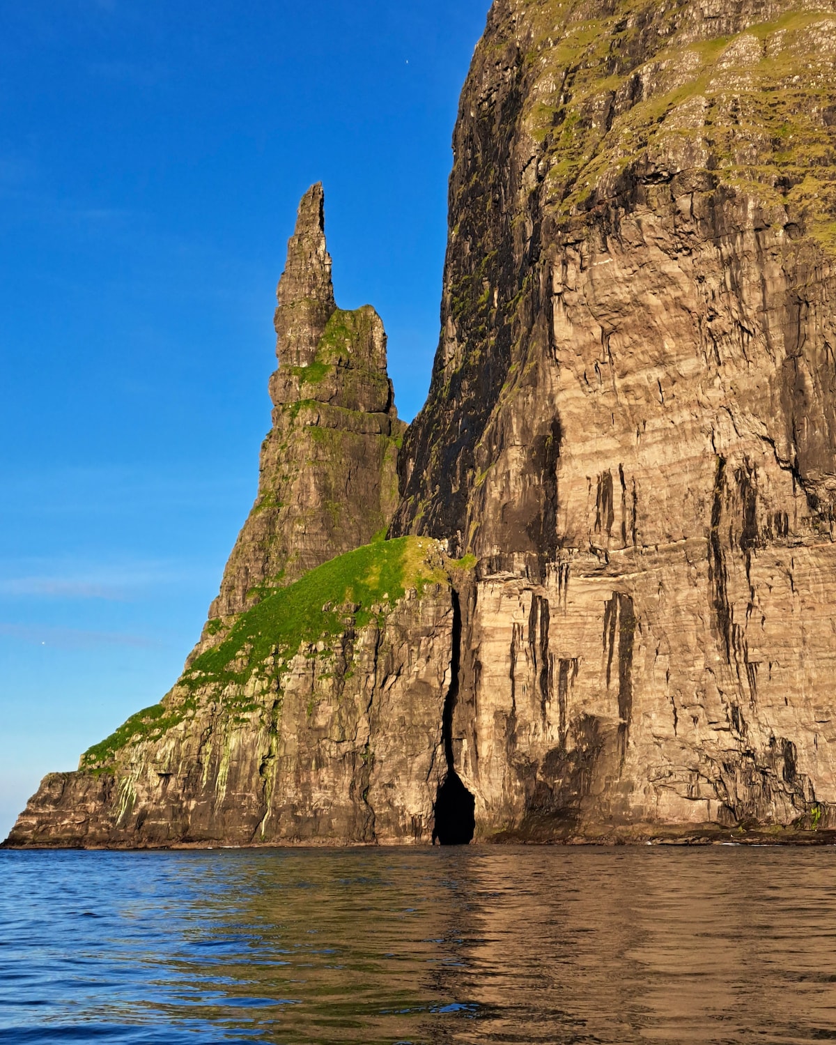 A towering rock formation rises sharply from the water, characterized by its rugged texture and steep cliffs. Lush green vegetation clings to the base, contrasting with the dark, weathered stone. The clear blue sky enhances the dramatic scenery reflected on the calm surface of the water.