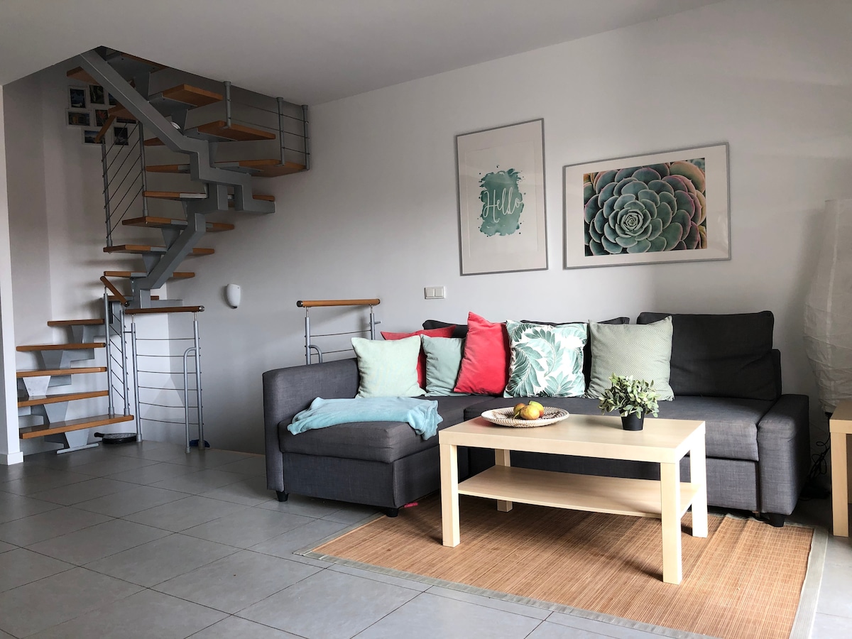 A modern living area is presented with a sectional sofa featuring various throw pillows in soft colors. A light wooden coffee table rests on a natural fiber rug, complemented by decorative plants. A staircase with metal and wood elements leads to the upper level.