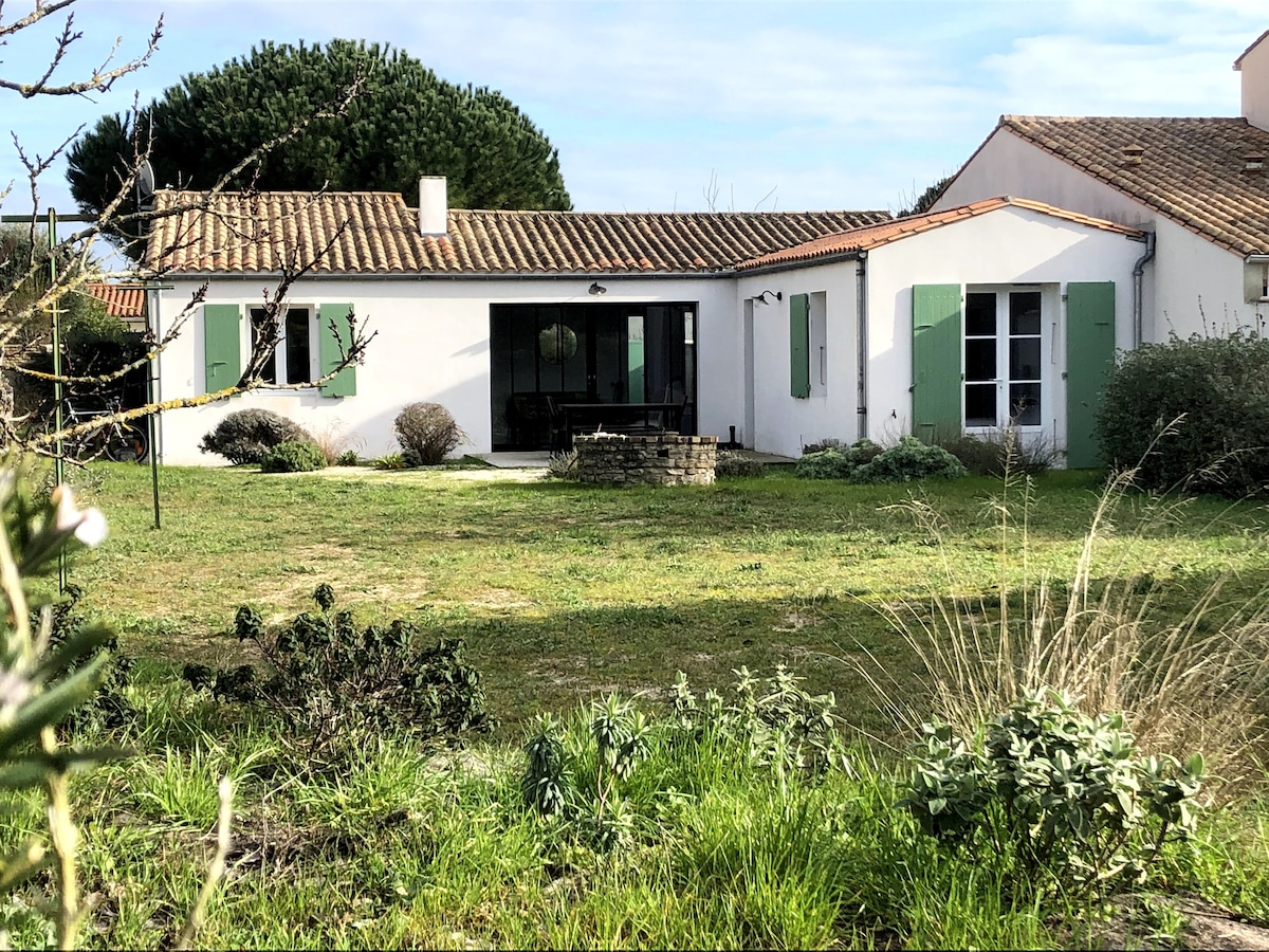 A single-story house is set amidst a spacious garden, featuring green shutters on white walls. A covered terrace is visible, providing outdoor seating. Lush grass and low shrubbery surround the structure, enhancing the sense of openness and proximity to nature.