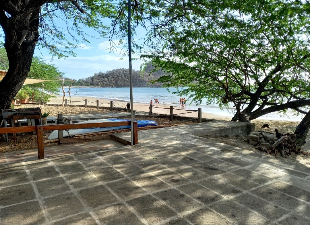 A spacious outdoor area is shown, featuring a paved surface extending towards a sandy beach. Lush trees provide shade, while the ocean view is complemented by a distant hillside. A kayak is resting nearby, and people can be seen enjoying the beach in the background.