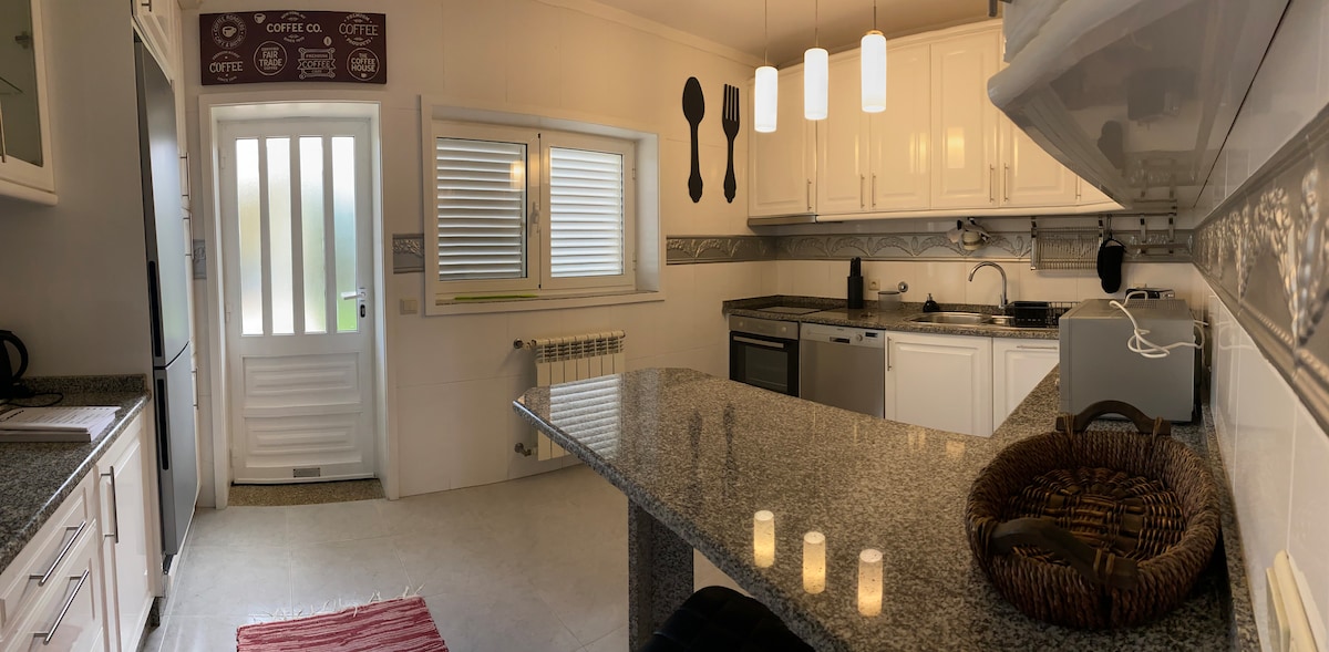 A modern kitchen features sleek white cabinetry and a granite countertop, highlighted by three pendant lights overhead. An array of essential appliances, including a built-in oven and stovetop, are visible, along with a cozy dining area that includes a woven basket. Natural light streams in through a window.
