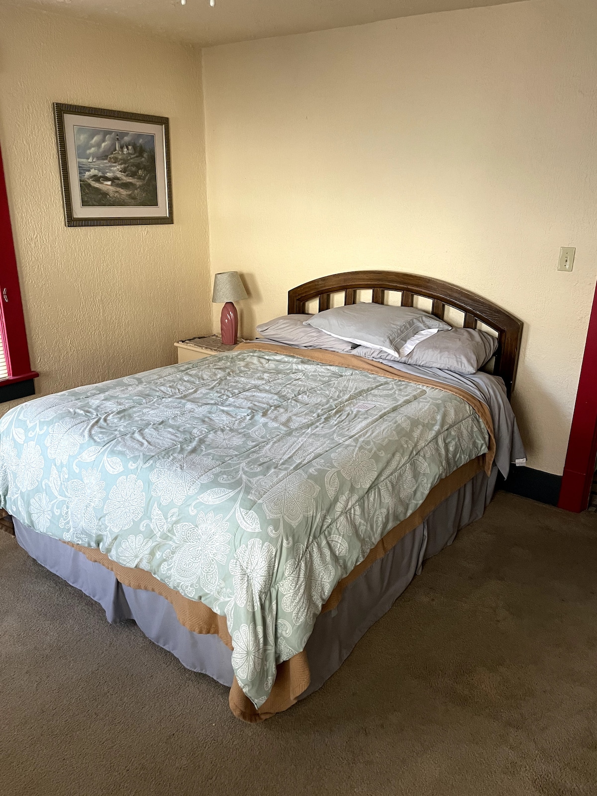 A cozy bedroom is highlighted by a spacious bed with a light-patterned comforter and neutral linens. A bedside lamp and a small nightstand flank the bed. Natural light enters through windows with red frames, accentuating the room's tranquil atmosphere.