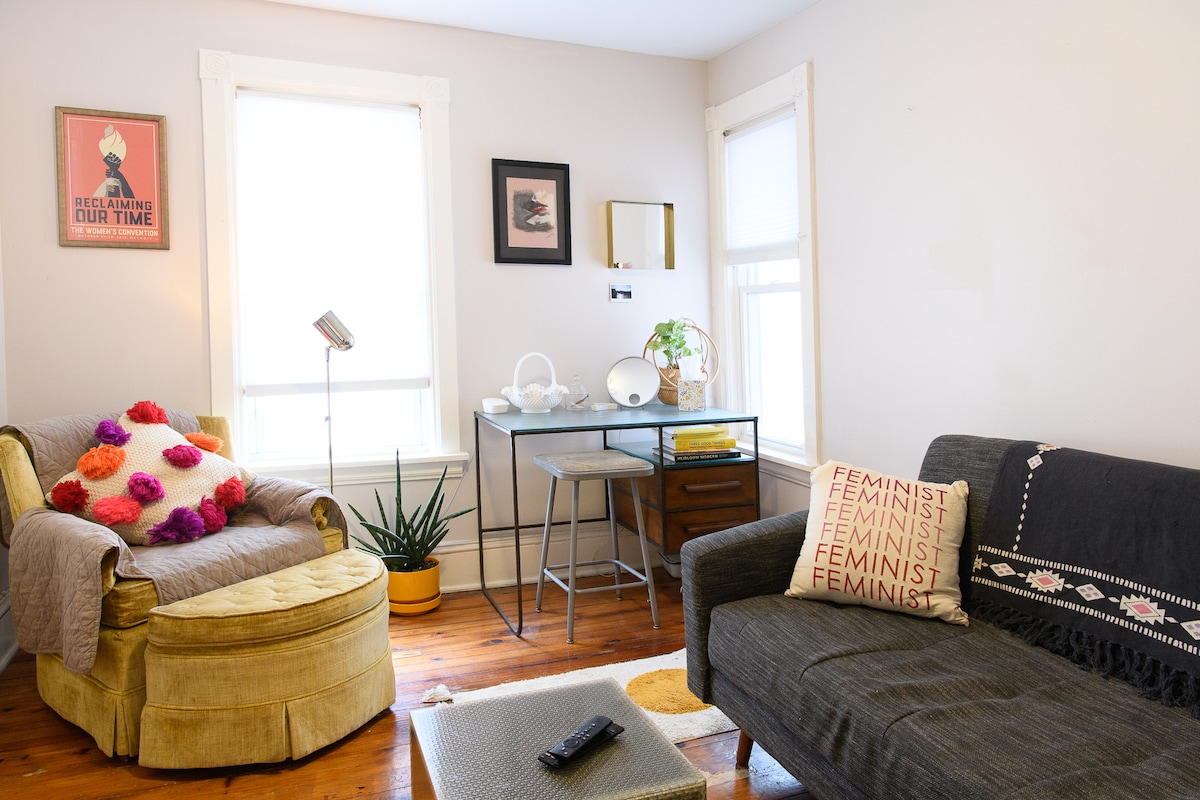 A living space is displayed featuring a vintage yellow armchair adorned with colorful pillows, alongside a dark gray couch. A small table with modern decor elements sits between them, while natural light enters through two windows, illuminating the warm wooden floor.