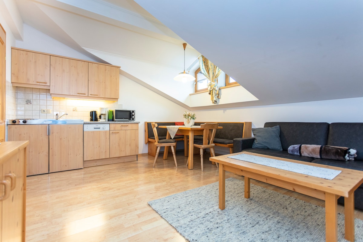 An open living area features wooden cabinetry and appliances, including a microwave and coffee maker. A dining table with chairs is positioned near a window, while a sofa is complemented by throw pillows. Natural light enhances the warm tones of the wooden floors.