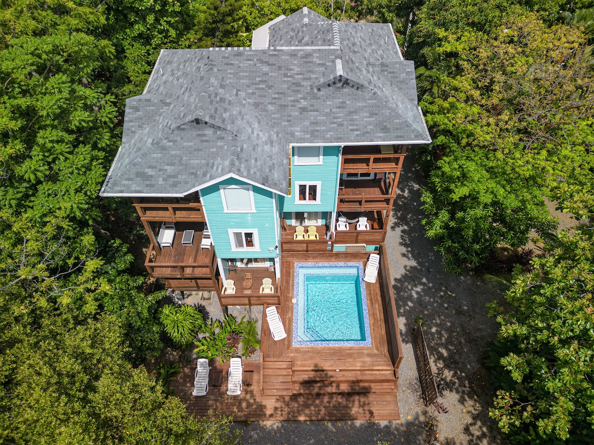 An aerial view captures a charming multi-level building surrounded by lush greenery. A private swimming pool with wooden decking can be seen below, along with outdoor seating areas. The building's exterior features a soft blue color, complemented by natural wood accents and multiple balconies.