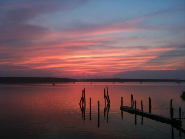 The image captures a serene waterfront sunset with vibrant pink and purple hues reflected on the water's surface. Silhouettes of mooring posts and distant boats are visible, enhancing the tranquil coastal atmosphere.