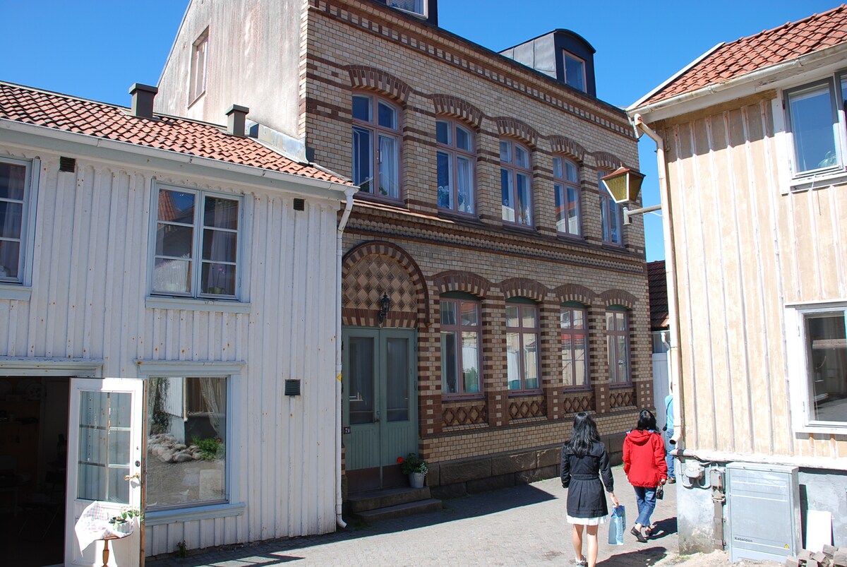 A charming building, characterized by its unique brick facade and decorative windows, stands at the center of the image. Two individuals walk towards the entrance, framed by neighboring wooden structures with traditional weathered exteriors. A clear blue sky complements the scene.