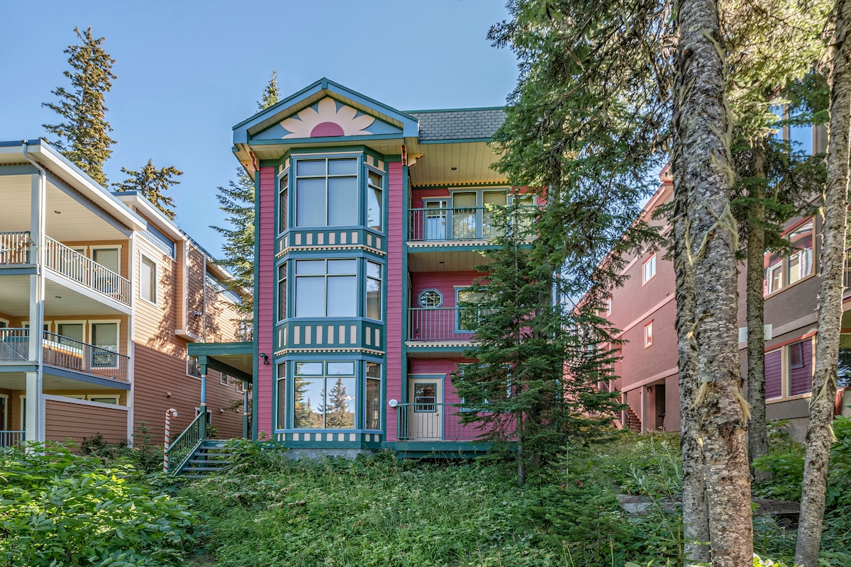 The exterior of the condo is framed by lush greenery and tall trees, featuring a distinctive architectural design with colorful accents. Large windows provide ample natural light, and a welcoming entrance is visible, inviting guests into the shared outdoor space.