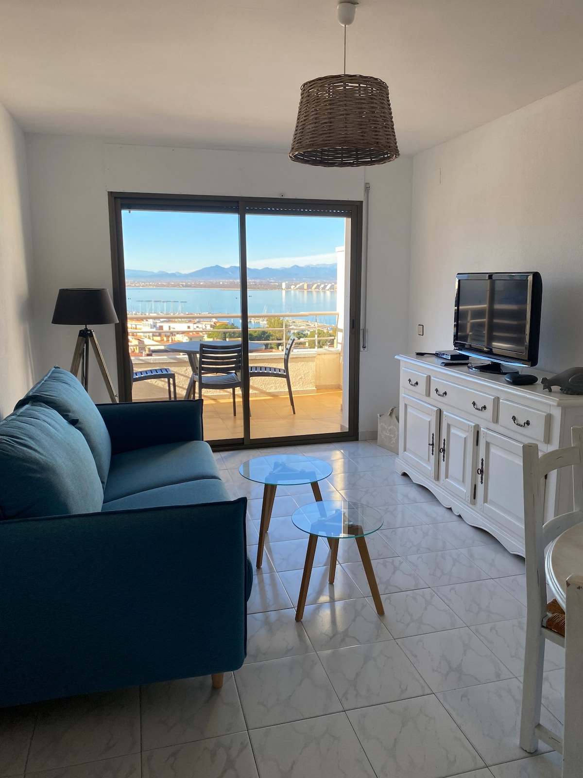 A living area features a blue sofa paired with two glass side tables. Large sliding doors provide access to a balcony with sea views. A television is situated on a white cabinet, with a lamp illuminating the space, complemented by natural light entering from the outdoor area.