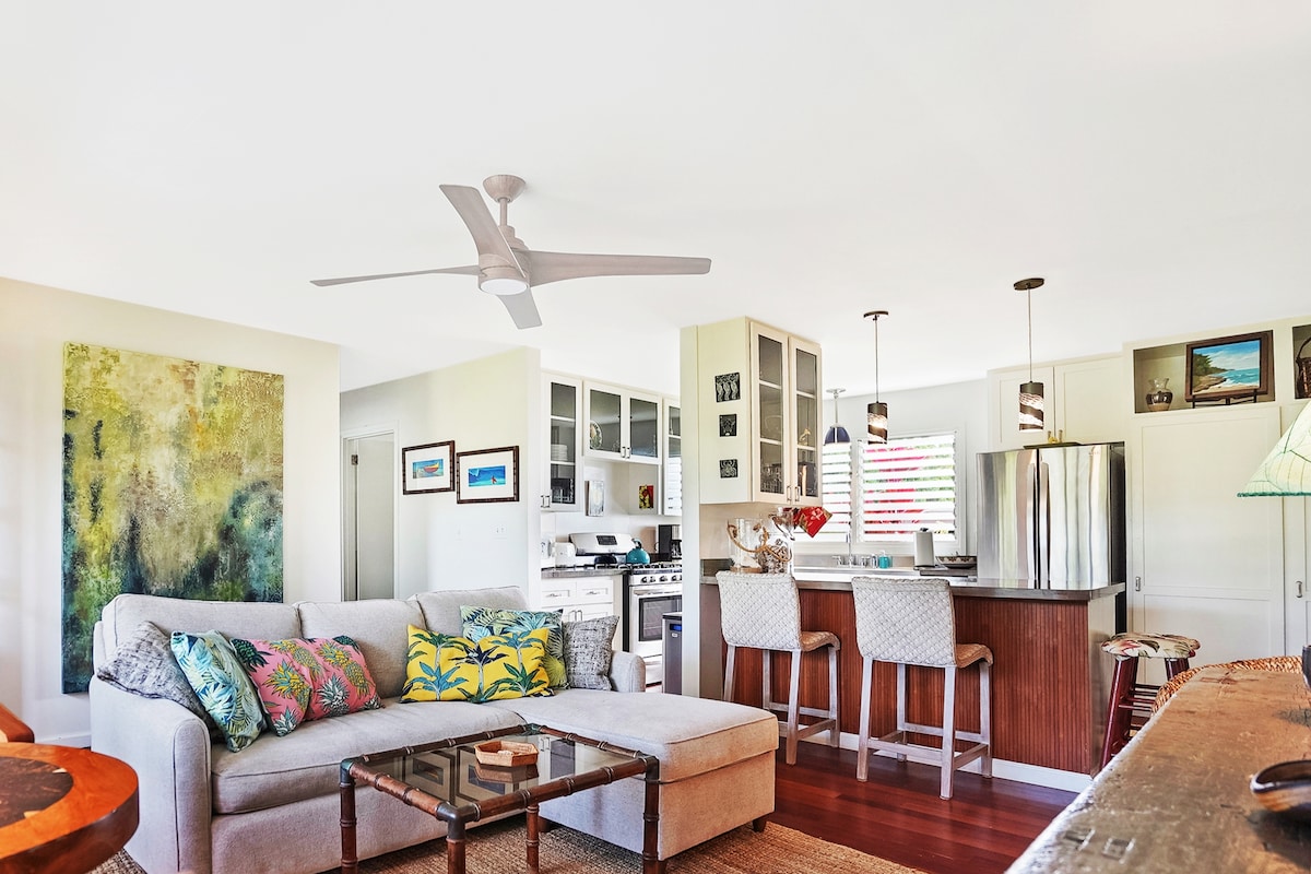 An open-concept living space features a light-colored sectional sofa adorned with colorful throw pillows. A modern kitchen with stainless steel appliances and a breakfast bar is visible in the background, complemented by vibrant artwork on the walls and natural light from large windows.