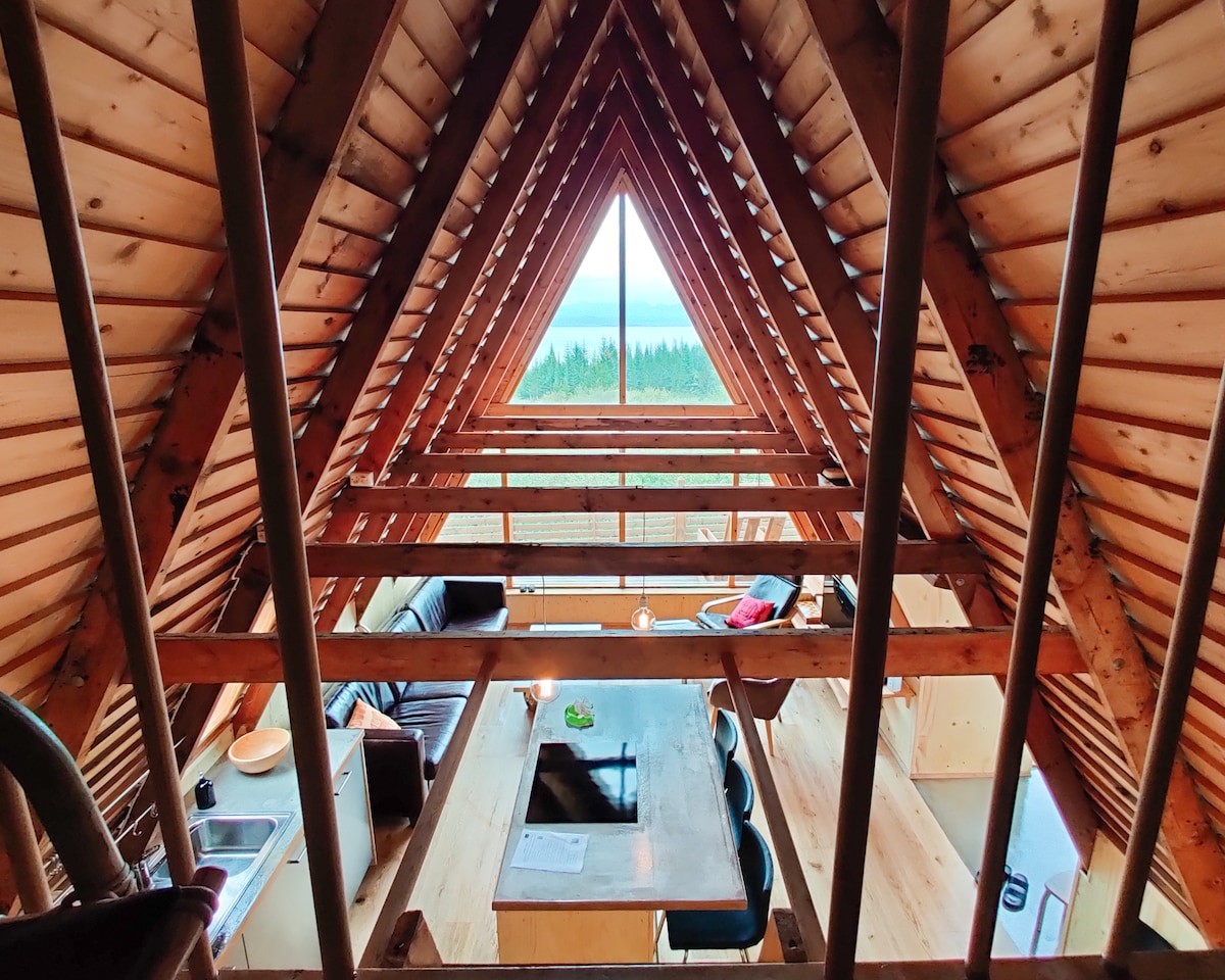 The interior features a vaulted wooden ceiling with exposed beams, creating an open and airy feel. A large triangular window offers a view of the surrounding landscape. A central dining table is visible, surrounded by black chairs, with a cozy seating area adorned with a dark sofa.