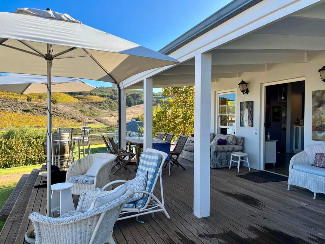 A spacious outdoor deck features comfortable seating arrangements with white wicker chairs and side tables. Shade is provided by large umbrellas. The view captures the rolling hills in the background, reflecting the natural landscape of the vineyard.