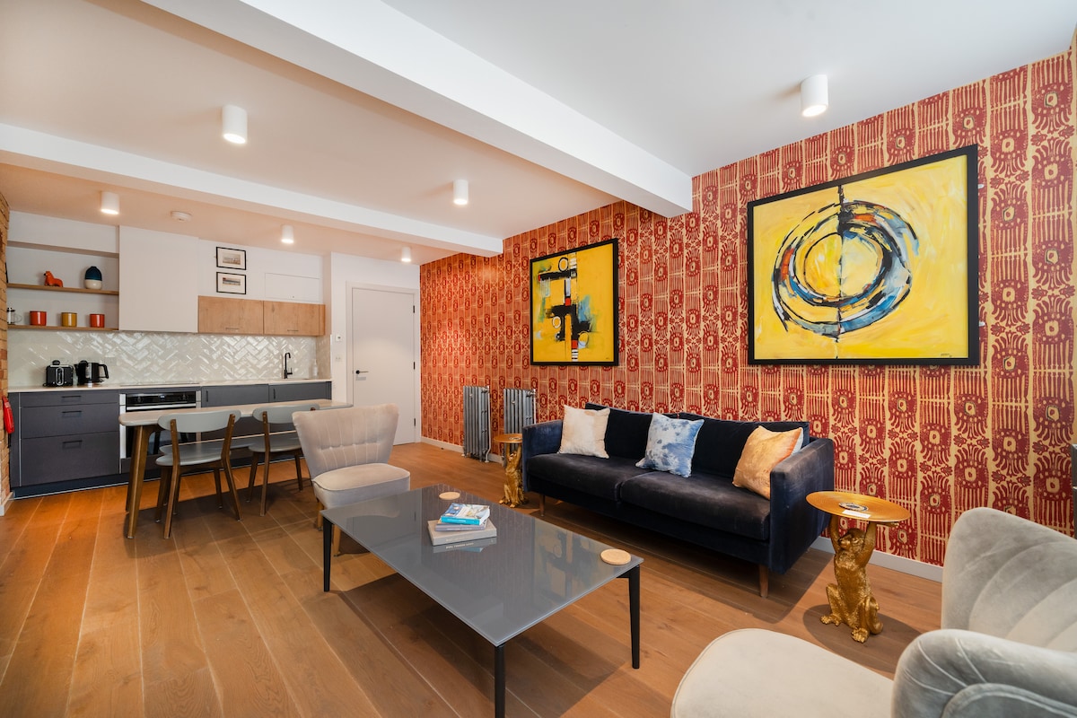 The open living area features vibrant art pieces on red patterned walls and a comfortable seating arrangement with a mix of sofas. A sleek glass coffee table sits in front of the seating, with a compact kitchen visible in the background and a light, airy ambiance created by ample ceiling lighting.