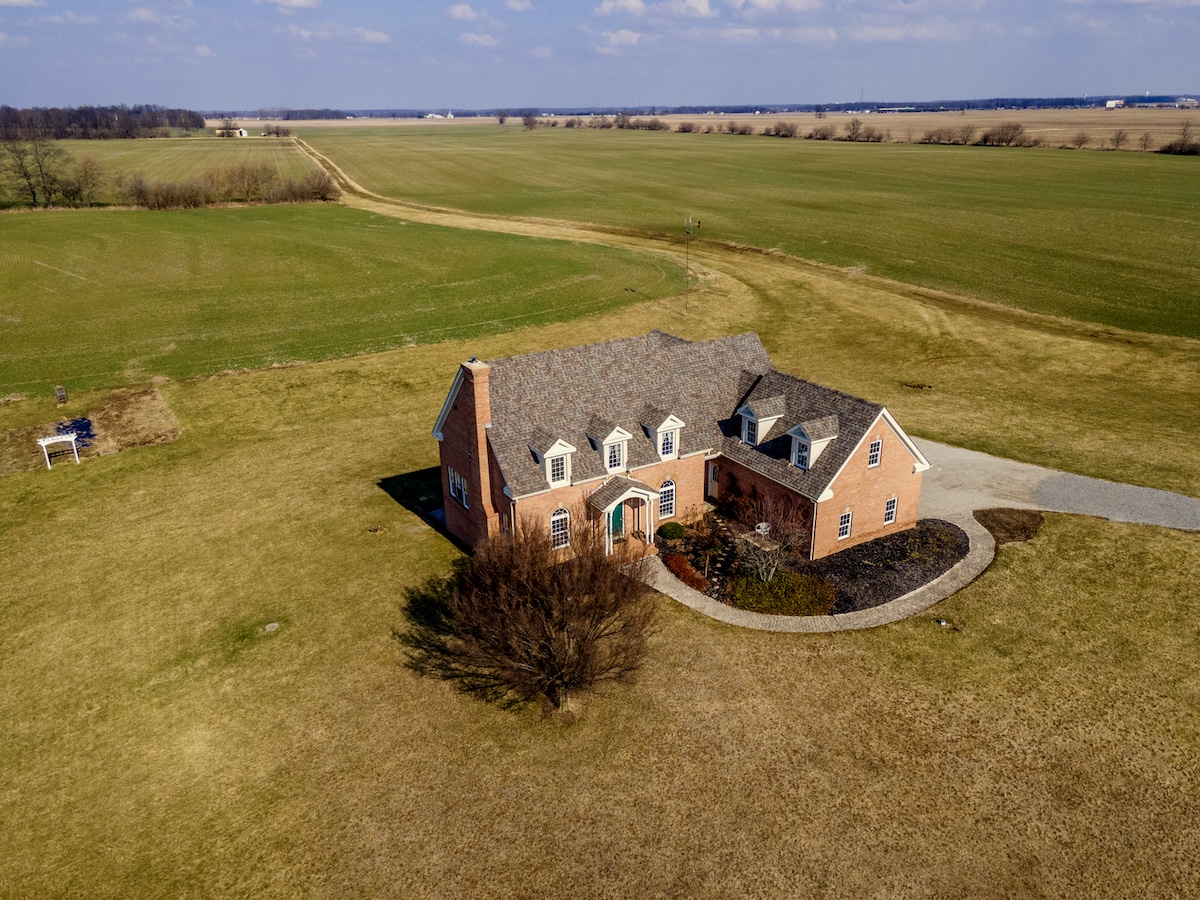 An aerial view of a spacious brick home surrounded by expansive green fields. The property features a circular driveway and a well-maintained landscape that complements the rural setting.