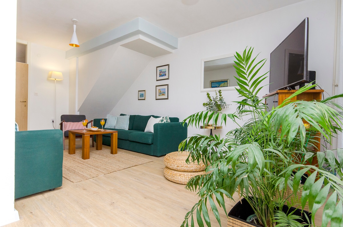 A spacious living room is filled with natural light, furnished with a green sofa and textured throw pillows. A wooden coffee table sits in the center, accompanied by a rug made of natural fibers. A television and potted greenery add to the inviting ambiance.