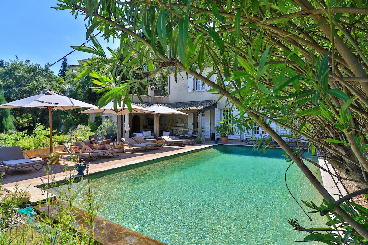 A natural swimming pool is surrounded by a stone patio with lounge chairs and shaded umbrellas. Lush greenery frames the scene, offering a sense of privacy and tranquility. The structure of the bastide is visible in the background, along with glimpses of the serene landscape.