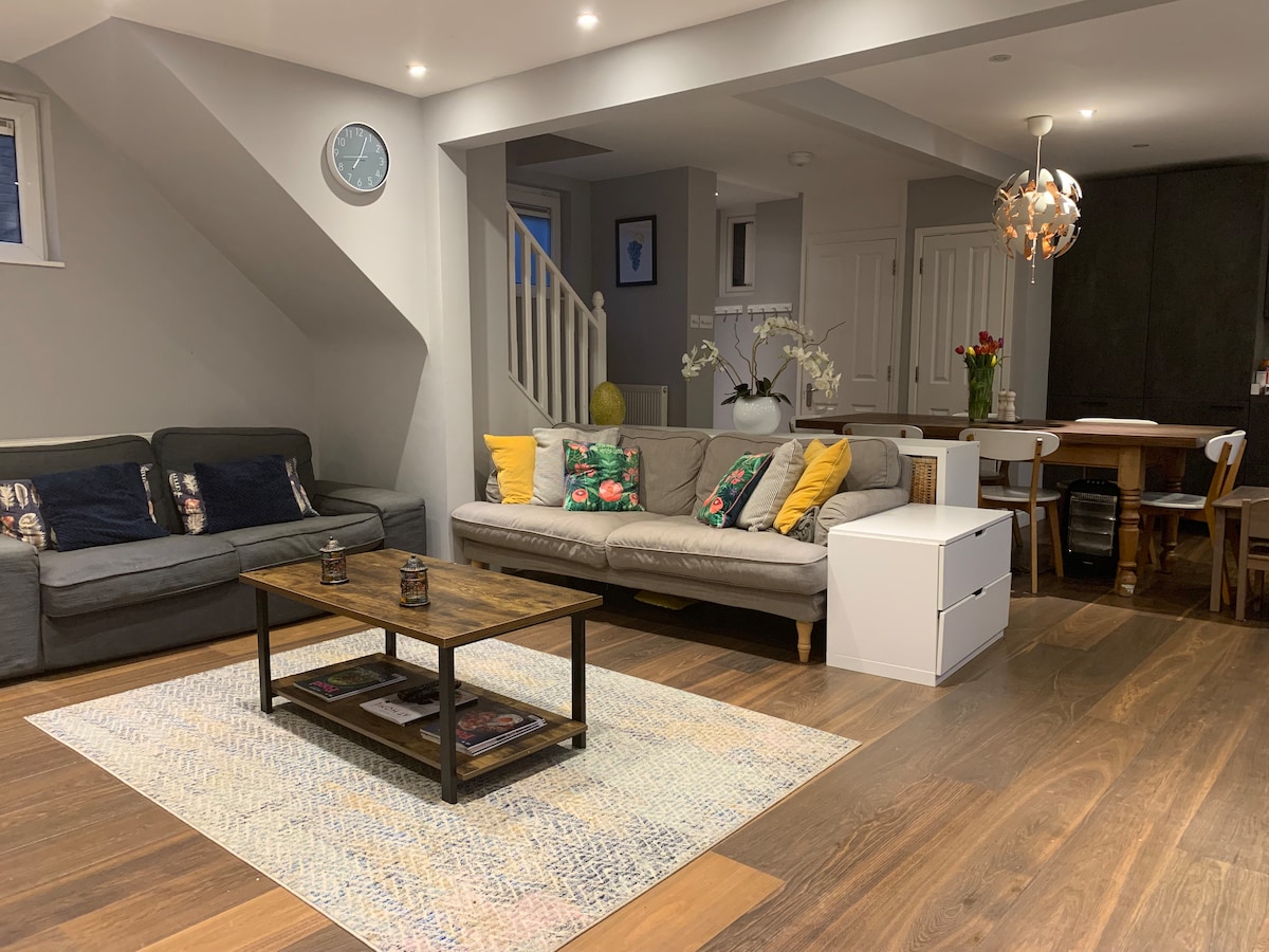 A cozy living area is furnished with two sofas, one adorned with colorful cushions. A wooden coffee table sits on a textured area rug. In the background, a dining area with a table and chairs is visible, along with modern kitchen cabinetry.